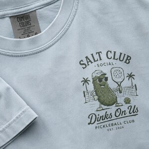 May include: A light blue t-shirt featuring a dark green graphic of a pickle wearing a cap and sunglasses, holding a pickleball paddle. The text reads SALT CLUB SOCIAL, Dinks On Us, PICKLEBALL CLUB, EST. 2026. The shirt has a Comfort Colors brand label.