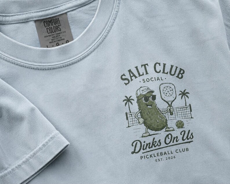 May include: A light blue t-shirt featuring a dark green graphic of a pickle wearing a cap and sunglasses, holding a pickleball paddle. The text reads SALT CLUB SOCIAL, Dinks On Us, PICKLEBALL CLUB, EST. 2026. The shirt has a Comfort Colors brand label.