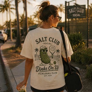 May include: A cream-colored short-sleeve t-shirt featuring a dark green graphic on the back. The design shows a pickle wearing a cap and sunglasses, holding a pickleball paddle, with the text SALT CLUB SOCIAL, Dinks On Us, and PICKLEBALL CLUB EST. 2026.