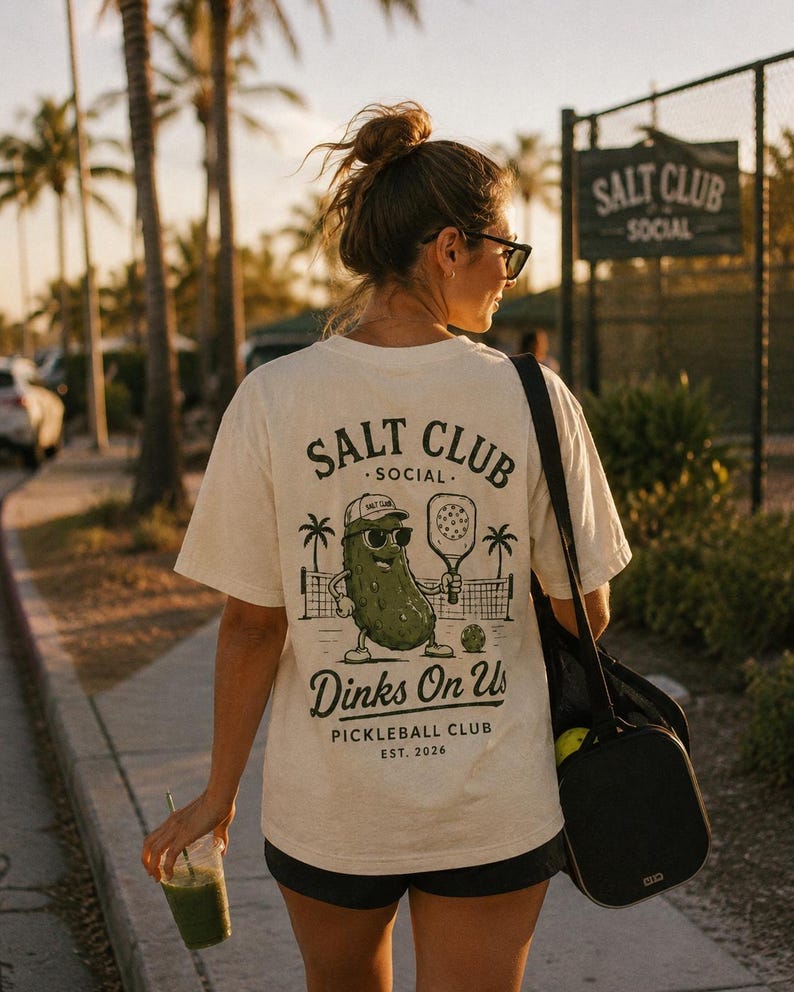 May include: A cream-colored short-sleeve t-shirt featuring a dark green graphic on the back. The design shows a pickle wearing a cap and sunglasses, holding a pickleball paddle, with the text SALT CLUB SOCIAL, Dinks On Us, and PICKLEBALL CLUB EST. 2026.