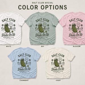 May include: Five short-sleeve t-shirts in white, bay, blossom, chambray, and ivory. Each shirt features a graphic of a pickle wearing sunglasses and a cap, holding a paddle, with the text SALT CLUB SOCIAL Dinks On Us PICKLEBALL CLUB EST. 2024.