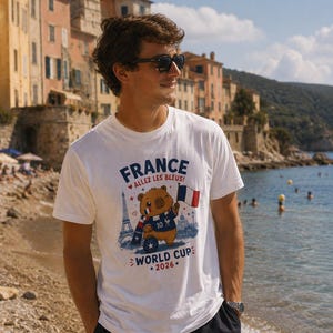 May include: A white short-sleeve t-shirt featuring a graphic of a brown bear wearing a scarf and jersey with the number 10, holding a French flag. The text reads FRANCE, ALLEZ LES BLEUS!, and WORLD CUP 2026, with an illustration of the Eiffel Tower.