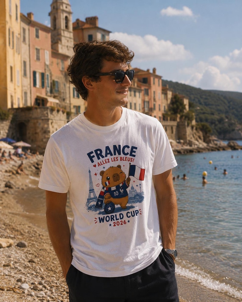 May include: A white short-sleeve t-shirt featuring a graphic of a brown bear wearing a scarf and jersey with the number 10, holding a French flag. The text reads FRANCE, ALLEZ LES BLEUS!, and WORLD CUP 2026, with an illustration of the Eiffel Tower.