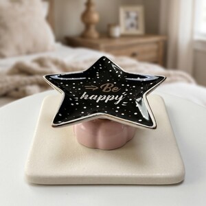 Könnte beinhalten: Eine schwarze, sternförmige Schmuckschale mit weißen Tupfen und goldenem Rand, die auf einem rosa, gewellten Sockel ruht. Auf der Schale steht in goldener Schrift "Be happy". Die Schale steht auf einem quadratischen, cremefarbenen Tablett.