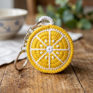Può includere: Un portachiavi a forma di fetta di limone all'uncinetto. Il portachiavi è un cerchio all'uncinetto giallo brillante con dettagli bianchi per imitare una fetta di limone. Una catena e un anello in metallo argentato sono attaccati in alto, con un anello all'uncinetto bianco.