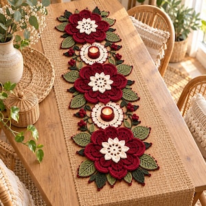 Puede incluir: Un camino de mesa largo y rectangular de tejido color canela, decorado con tres grandes flores de ganchillo rojo oscuro con centros color crema, rodeadas de hojas de ganchillo verdes, pequeñas bayas rojas y dos posavelas circulares color crema.