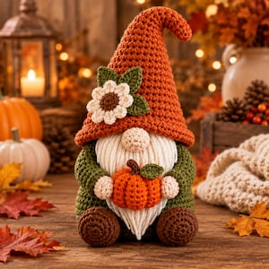 May include: A crocheted gnome figurine featuring a tall, textured orange hat adorned with a white and brown flower and green leaves. The gnome has a cream-colored beard, dark green sleeves, brown feet, and holds a small orange pumpkin.
