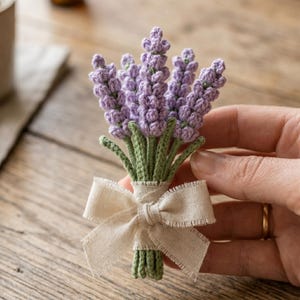 May include: A handmade crochet lavender bouquet with purple flower heads, green stems, and a beige ribbon bow. The bouquet is held in a hand, highlighting the intricate yarn details. The theme is floral and handcrafted.