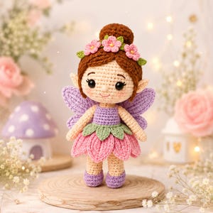 Pode incluir: Uma boneca fada de crochê com cabelo castanho num coque, adornada com flores cor-de-rosa e folhas verdes. A boneca apresenta asas roxas, um corpete roxo, uma saia de pétalas cor-de-rosa e sapatos roxos, com olhos pretos e um sorriso.