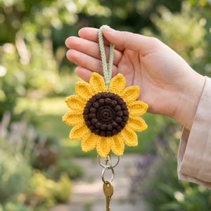 May include: A crocheted sunflower keychain with a yellow and brown flower design. The keychain has a green loop and holds a set of keys. The sunflower is held in front of a blurred green background.