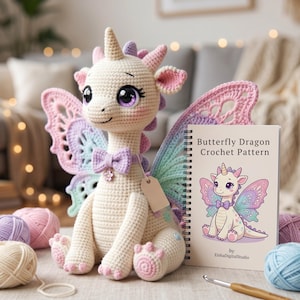 May include: A cream-coloured crocheted dragon with butterfly wings, a bow tie, and a small flower pendant. The dragon has pink, purple, and blue accents. A book titled "Butterfly Dragon Crochet Pattern" is next to the dragon.