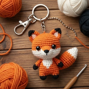 May include: A crocheted fox keychain in orange, white, and black. The fox has large black eyes and a white chest and tail tip. It is attached to a silver keychain with a lobster clasp. Yarn balls and a crochet hook are in the background.