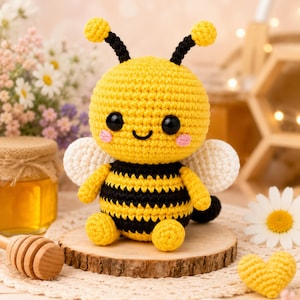 May include: A crocheted bee plush toy featuring a round yellow head with black antennae, large black eyes, and pink cheeks. The body has yellow and black horizontal stripes, small white wings, and yellow limbs. It sits on a round wooden base.