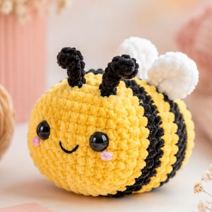 May include: A handmade, plush crochet bee with a smiling face. The bee features a yellow body with black stripes, black antennae, and white wings. The bee's cheeks are pink, and it has large, shiny black eyes.