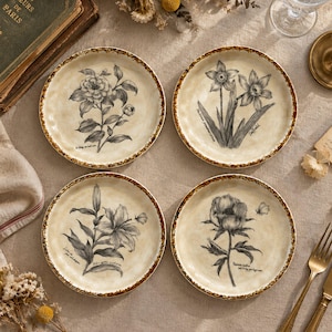 May include: A set of four round ceramic plates with a cream-coloured base and a mottled brown rim. Each plate features a distinct black and white botanical illustration of a flower, including a rose, daffodils, a lily, and a peony, with script text.