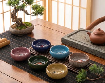 Op de afbeelding: Een verzameling kleine, kleurrijke keramische theekommen in roze, blauw, groen en geel, gerangschikt op een houten tafel met een theeset en een bonsai-boom. De kommen hebben een glanzende afwerking.