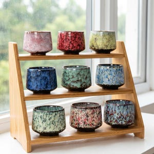 May include: Nine ceramic teacups in various colors, including red, blue, and green, are displayed on a tiered bamboo shelf. Each cup has a textured surface and a dark brown base. The cups are arranged in three rows of three.