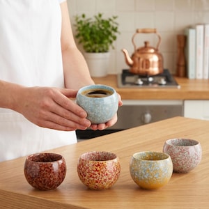 May include: A set of five round ceramic cups featuring a speckled, multi-colored glaze. One light blue cup is held, while four others in dark red, orange-red, yellow-blue, and pink-grey sit on a wooden surface, each with a smooth, rounded shape.