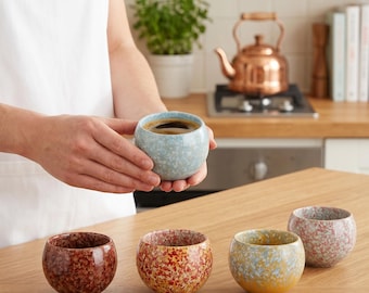 Op de afbeelding: Een set van vijf ronde keramische bekers met een gespikkeld, veelkleurig glazuur. Eén lichtblauwe beker wordt vastgehouden, terwijl vier andere in donkerrood, oranje-rood, geel-blauw en roze-grijs op een houten oppervlak staan.