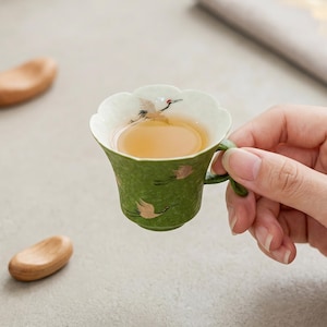 May include: A small, green ceramic teacup with a scalloped rim and a handle. The exterior and interior feature painted illustrations of flying cranes with gold accents. The cup is filled with a light amber-coloured tea, showcasing a delicate design.