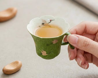 Op de afbeelding: Een kleine, groene keramische theekop met een geschulpte rand en een oortje. De buiten- en binnenkant zijn voorzien van geschilderde afbeeldingen van vliegende kraanvogels met gouden accenten. De kop is gevuld met licht amberkleurige thee.