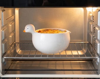 Op de afbeelding: Een witte keramische kom in de vorm van een eend met een oranje snavel, gevuld met een goudbruin gebakken gerecht. De kom staat op het metalen rooster in een roestvrijstalen oven met een verlichte binnenkant en een glazen deur.