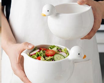 Op de afbeelding: Twee witte keramische kommen, elk voorzien van een gevormd handvat in de vorm van een eendenkop met een gele snavel en zwarte ogen. Eén kom is leeg, terwijl de andere gevuld is met een verse salade van komkommerschijfjes, tomaten en sla.