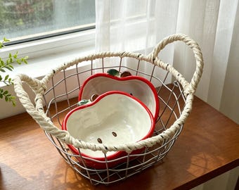 Op de afbeelding: Een draadmand met twee met touw omwikkelde handvatten bevat twee keramische kommen in de vorm van appels. De kommen hebben een glanzend witte binnenkant, een felrode buitenrand en een detail van een groen blaadje en bruin steeltje.