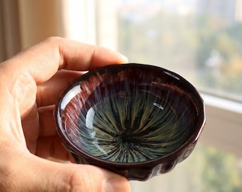 Op de afbeelding: Een kleine, handgemaakte keramische theekom met een unieke glazuur. De kom heeft een donkerbruine buitenkant en een wervelend interieurontwerp met tinten groen, blauw en goud. De rand heeft een golvende textuur.