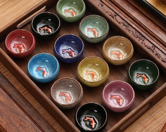 Op de afbeelding: Een set van twaalf kleine, kleurrijke keramische theekopjes, gerangschikt in een houten dienblad. Elke kop heeft een gedetailleerd, driedimensionaal goudvisontwerp op de bodem. De kopjes zijn in verschillende kleuren, waaronder rood, blauw, groen en geel.