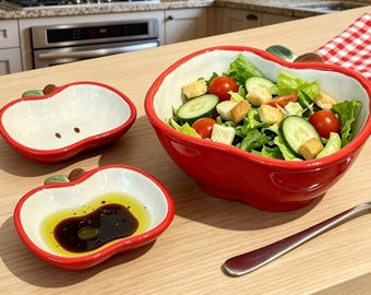 Op de afbeelding: Een set van drie appelvormige keramische schalen met een glanzende rode buitenkant en witte binnenkant. Een grote kom bevat een salade met sla, komkommer, tomaten en croutons. Twee kleinere schaaltjes bevatten olijfolie en balsamicoazijn.