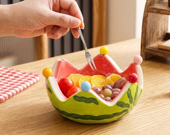 Op de afbeelding: Een keramische kom in de vorm van een watermeloenpartje met een geschulpte rand voorzien van kleine, kleurrijke bolletjes op elk punt. De buitenkant is groen met donkergroene strepen en de binnenkant is roze. Het bevat kerstomaatjes en druiven.