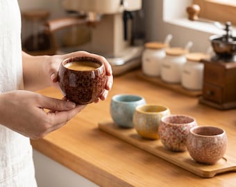Op de afbeelding: Een set van vijf kleine, ronde keramische kopjes met een gespikkeld, getextureerd glazuur. Eén donkerbruin kopje wordt op de voorgrond vastgehouden, terwijl vier andere in lichtblauw, geel, roze en mauve op een houten dienblad staan.