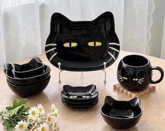 Op de afbeelding: Een zwarte keramische serviesset met katvormige ontwerpen, gele ogen en witte snorharen. De set bevat een groot bord in de vorm van een kat, een mok, verschillende kleine kommen en een stapel kleine vierkante schaaltjes met glanzende afwerking.