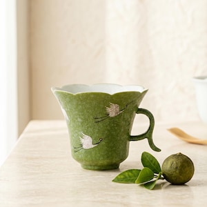 May include: A green ceramic mug with a scalloped rim and a textured finish, featuring white crane illustrations with black and red details. The mug has a curved handle and a white interior, accompanied by a small green citrus fruit with leaves.