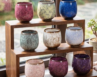 Op de afbeelding: Een verzameling van negen keramische theekopjes gerangschikt op een houten standaard met meerdere niveaus. Elke kop heeft een unieke craquelé glazuurafwerking in diverse kleuren, waaronder roze, geel, blauw, groen, wit, lichtblauw, lichtroze en paars.