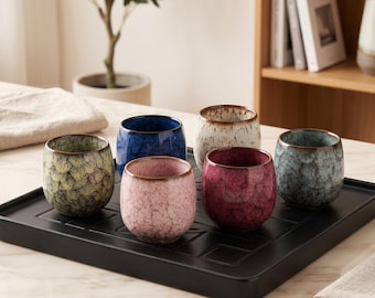 Op de afbeelding: Een set van zes kleine keramische bekers met een ronde vorm en een craquelé glazuurafwerking in diverse kleuren zoals groen, blauw, wit, roze, donkerrood en groenblauw, geplaatst op een zwart rechthoekig dienblad met raster.
