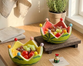 Op de afbeelding: Twee keramische kommen in de vorm van watermeloenpartjes met geschulpte randen bekroond met kleine kleurrijke bolletjes. De ene kom is geel en groen, de andere roze en groen. Beide bevatten diverse stukjes fruit zoals druiven en tomaten.