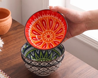 Op de afbeelding: Een stapel van drie keramische kommen. De bovenste kom heeft een levendig rood en oranje zonnestraalpatroon. De twee kommen eronder tonen een groen en wit geometrisch bladpatroon aan de binnenkant en een zwart en wit vierpaspatroon aan de buitenkant.
