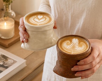 Op de afbeelding: Twee keramische mokken in de vorm van verfrommelde papieren zakken, elk gevuld met een latte met ingewikkelde schuimkunst. De ene mok is crèmewit, terwijl de andere een diepe, getextureerde bruine kleur heeft met een brede rand.