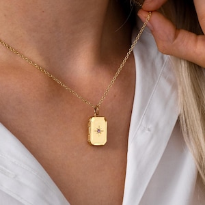 May include: A gold-toned rectangular locket pendant suspended on a delicate matching chain. The locket features a centred starburst engraving with a small purple gemstone inlay, set against a smooth, polished metallic surface.