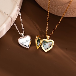 Könnte beinhalten: Eine silberfarbene Herzmedaillon-Halskette mit der Gravur Sophia und eine goldfarbene Herzmedaillon-Halskette an einer Kette, die geöffnet ist und ein kleines Babyfoto im Inneren zeigt. Beide Halsketten haben feine Metallgliederketten.