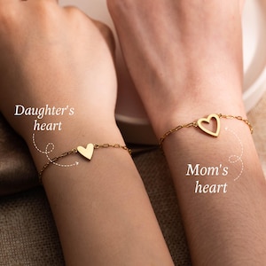 Puede incluir: Dos pulseras doradas con forma de corazón, una maciza y otra abierta, se muestran en las muñecas. Las pulseras están conectadas por una cadena. El texto de la imagen dice "Daughter's heart" y "Mom's heart".
