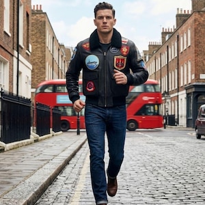 May include: A black leather bomber jacket with patches, worn with blue jeans and brown boots. The jacket has a black fur collar. A red double-decker bus is in the background, along with a black taxi cab and brick buildings.
