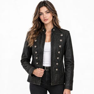 Könnte beinhalten: Eine schwarze Jacke im Militärstil mit strukturierter Silhouette, hohem Kragen und zwei vertikalen Reihen silberfarbener Metallknöpfe auf der Vorderseite. Die Jacke wird über einem schlichten weißen Oberteil und zu einer schwarzen Jeans getragen.