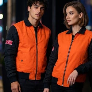 Puede incluir: Una chaqueta bomber con cremallera, torso naranja brillante y mangas, cuello y dobladillo negros en contraste. La manga derecha muestra un parche rectangular con el texto GOKU, mientras que la manga izquierda tiene un parche circular con el número 59.