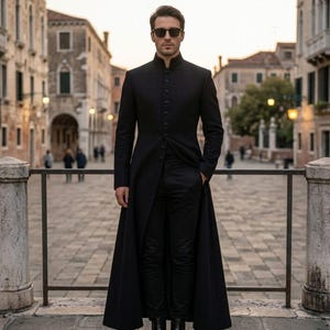May include: A person in a long, black coat with a mandarin collar and black trousers. The coat is buttoned up and reaches the ankles. They are wearing sunglasses and standing in a European city, possibly Venice.
