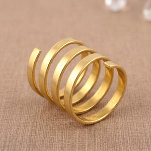 Puede incluir: Un anillo dorado con un diseño en espiral. El anillo presenta cinco bandas conectadas que se enrollan alrededor del dedo. El anillo está hecho de un material metálico brillante y está sobre un fondo neutro.