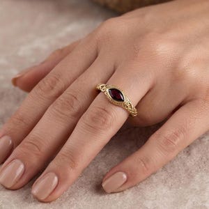 Könnte beinhalten: Ein Goldring mit einem tiefroten Edelstein im Marquise-Schliff, gefasst in eine Ringschiene mit filigranen Metallarbeiten. Die Ringschiene zeigt ein strukturiertes, verziertes Muster an den Seiten und rahmt den zentralen Stein in Gold ein.