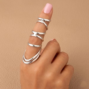 May include: A set of five silver-toned rings worn on a single finger. The collection includes two open-ended rings with a cross-bar design at the top, one simple smooth band, and two curved, layered bands positioned at the base of the finger.